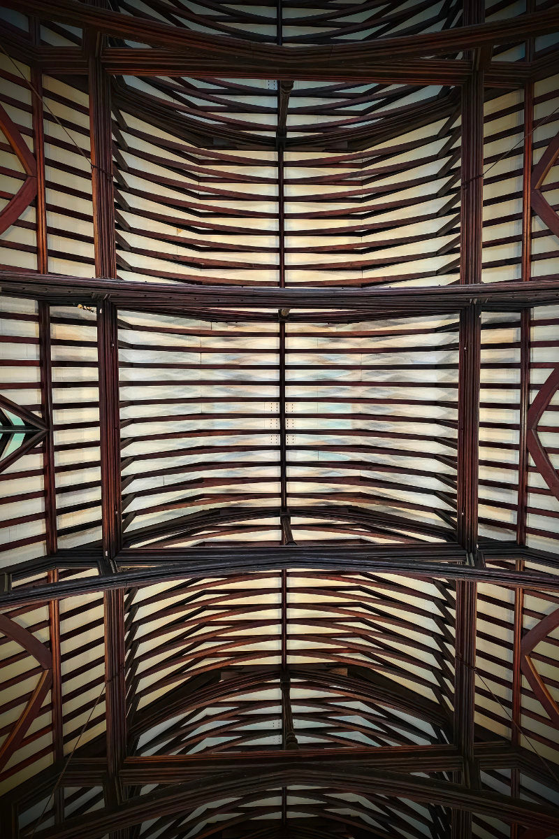 An upward view of a large arched ceiling with a repeating pattern of dark brown wooden beams and light-colored panels.