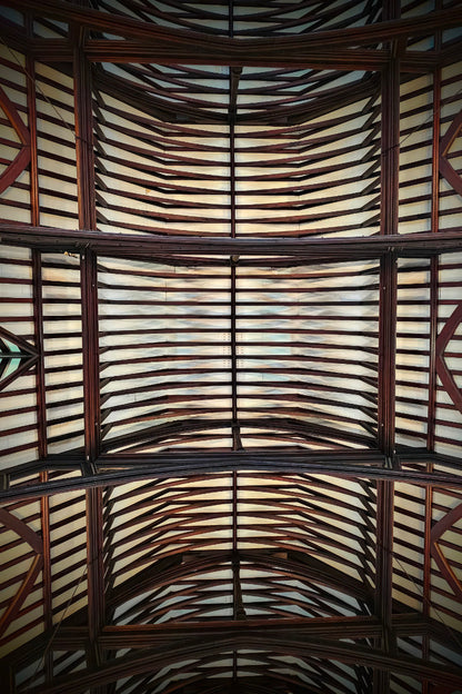 An upward view of a large arched ceiling with a repeating pattern of dark brown wooden beams and light-colored panels.