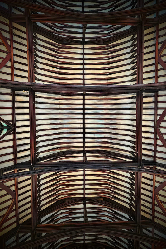 An upward view of a large arched ceiling with a repeating pattern of dark brown wooden beams and light-colored panels.