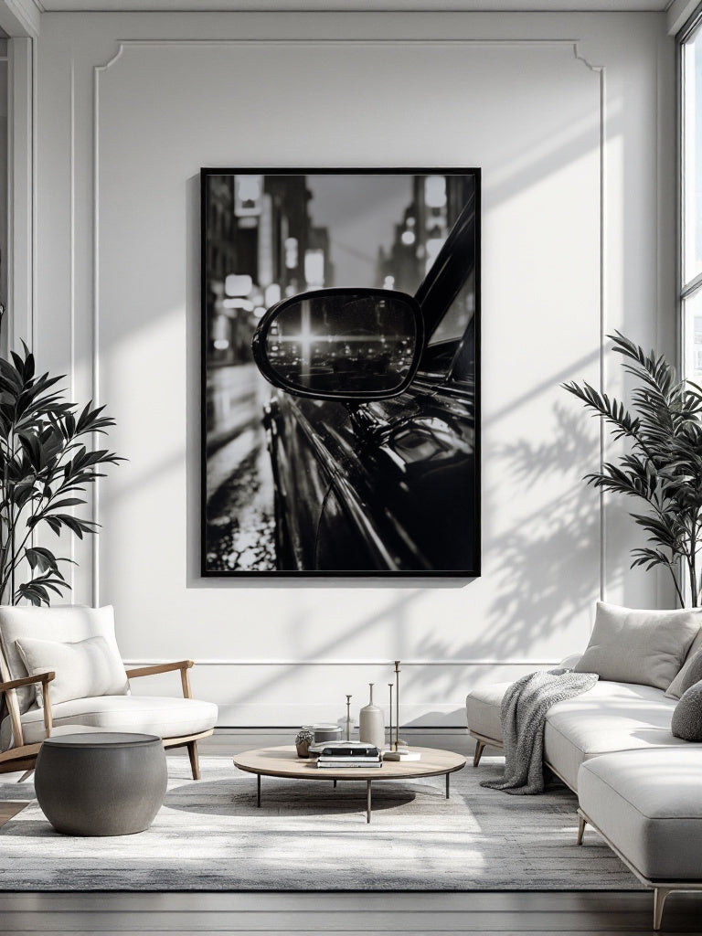 A large black and white framed wall art print depicting a city street scene with a car mirror, displayed in a modern living room setting with a sofa, armchair, coffee table, and potted plants.