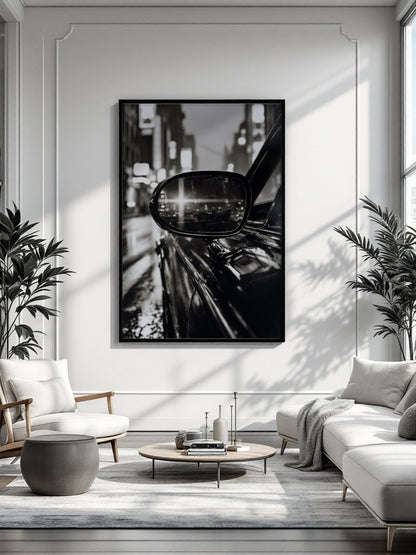A large black and white framed wall art print depicting a city street scene with a car mirror, displayed in a modern living room setting with a sofa, armchair, coffee table, and potted plants.