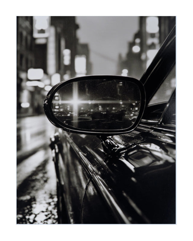 Black and white fine art photograph of a car side mirror reflecting a blurred city street at night, presented with a white border.