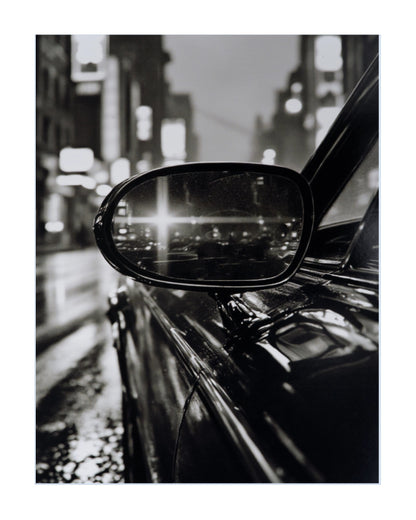 Black and white fine art photograph of a car side mirror reflecting a blurred city street at night, presented with a white border.