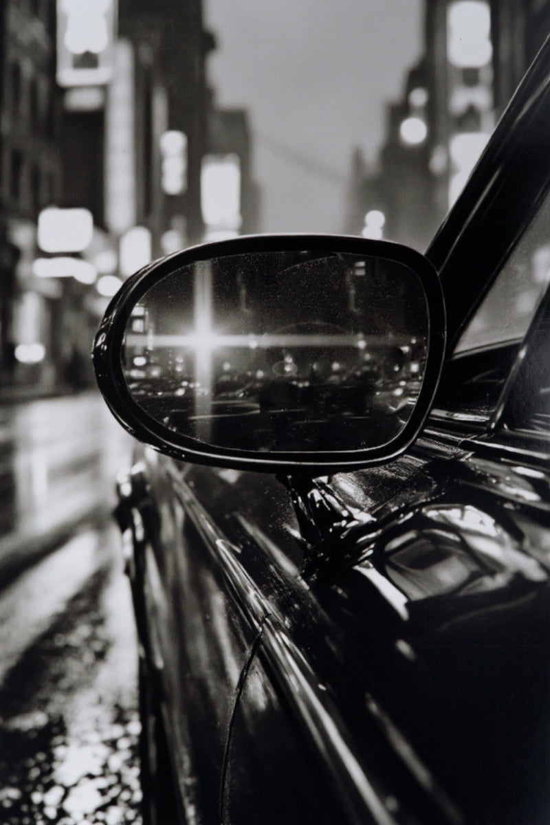Black and white close-up photograph of a car side mirror reflecting a blurred city street at night.