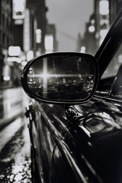 Black and white close-up photograph of a car side mirror reflecting a blurred city street at night.