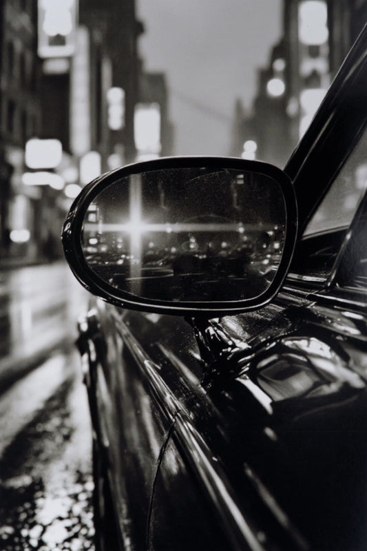 Black and white close-up photograph of a car side mirror reflecting a blurred city street at night.
