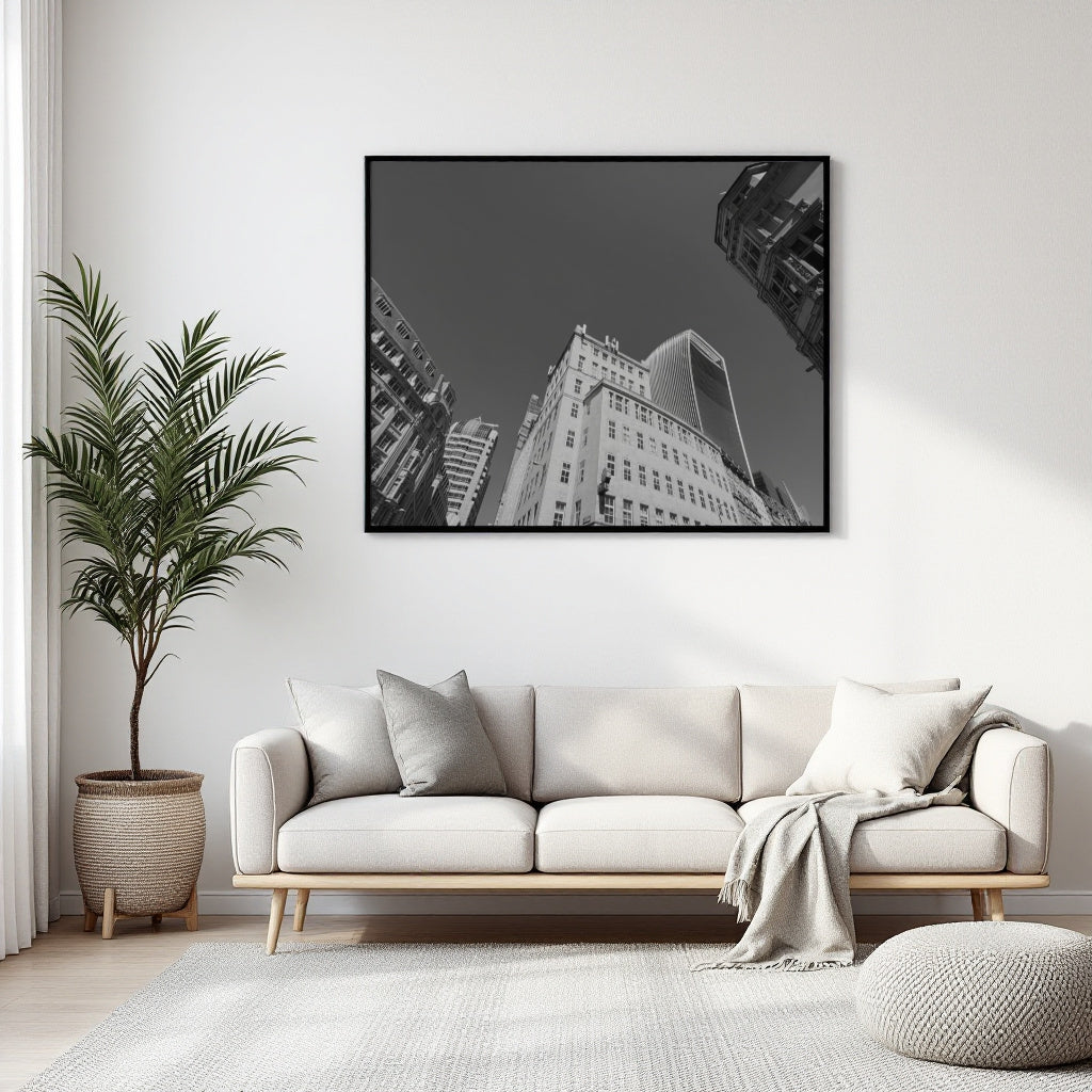 A black and white framed wall art print depicting a low-angle view of skyscrapers, displayed above a modern light beige sofa in a bright, contemporary living room.