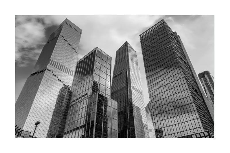 Black and white photograph of modern glass skyscrapers viewed from a low angle, with dramatic reflections in the windows against a cloudy sky.