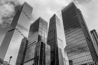 Black and white photograph of modern glass skyscrapers in a city, shown from a low angle looking up at the buildings against a cloudy sky.