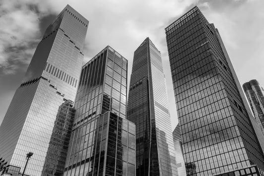 Black and white photograph of modern glass skyscrapers in a city, shown from a low angle looking up at the buildings against a cloudy sky.