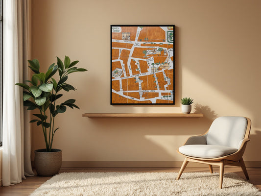 A framed art print featuring an orange and white street map design hangs on a beige wall in a modern living room setting above a wooden floating shelf, next to a green potted plant and a light wood armchair.