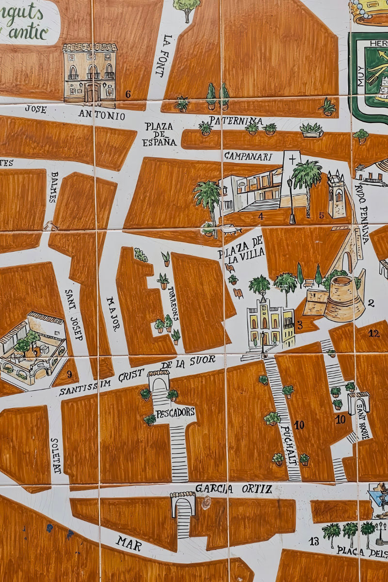 A close-up of a hand-painted ceramic tile map depicting the historic streets of Alicante, Spain, with white roads, brown buildings, and green accents.