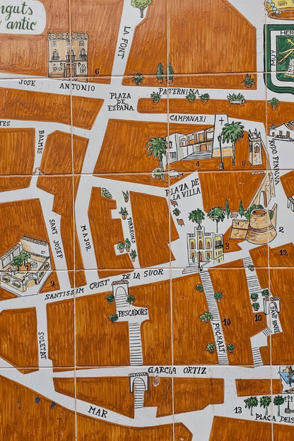 A close-up of a hand-painted ceramic tile map depicting the historic streets of Alicante, Spain, with white roads, brown buildings, and green accents.