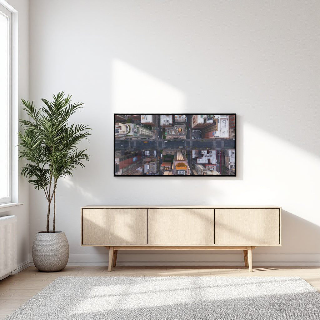 A framed wall art print showing a collage of street scenes, displayed above a light wood sideboard in a bright, modern room with a potted palm and window light.