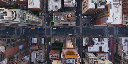 Aerial overhead view of a dense urban cityscape with intersecting streets, tall buildings, rooftops, and small vehicles, resembling a city map or print.