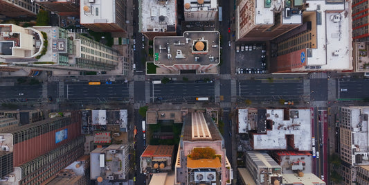 Aerial overhead view of a dense urban cityscape with intersecting streets, tall buildings, rooftops, and small vehicles, resembling a city map or print.