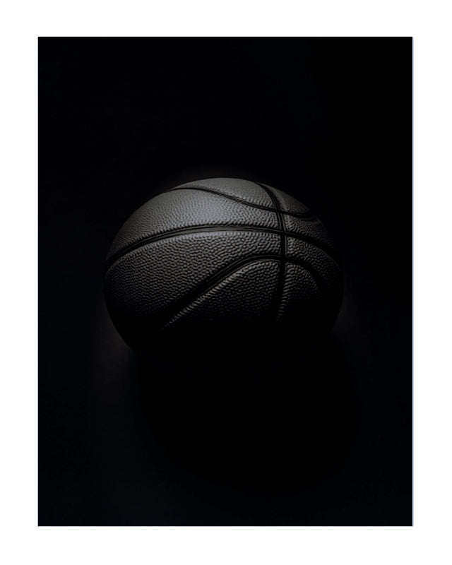 A black basketball with visible pebbled texture and dark painted seams, photographed against a very dark black background with dramatic low-key lighting.