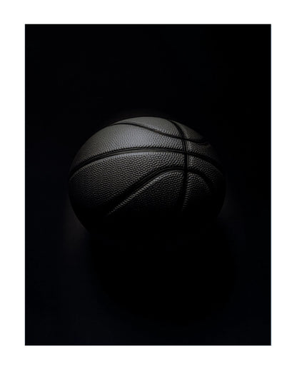 A black basketball with visible pebbled texture and dark painted seams, photographed against a very dark black background with dramatic low-key lighting.