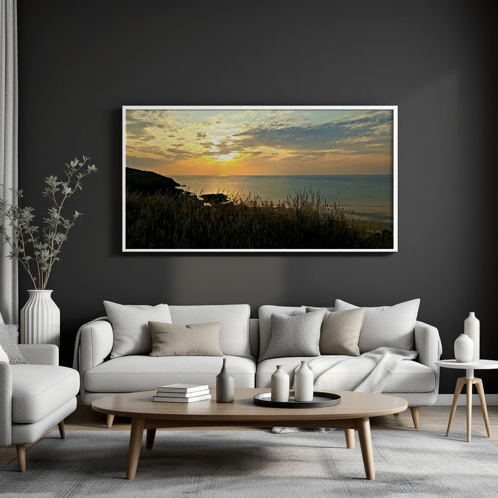 A framed horizontal landscape print of a sunset over the sea with grassy foreground, displayed on a dark charcoal wall above a modern white sofa in a styled living room.