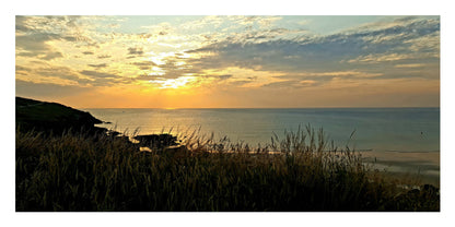 A scenic landscape photograph of a coastal sunset, featuring a dark silhouetted grassy foreground, rocky shoreline, calm sea, and a glowing yellow-orange sun beneath a cloudy sky.