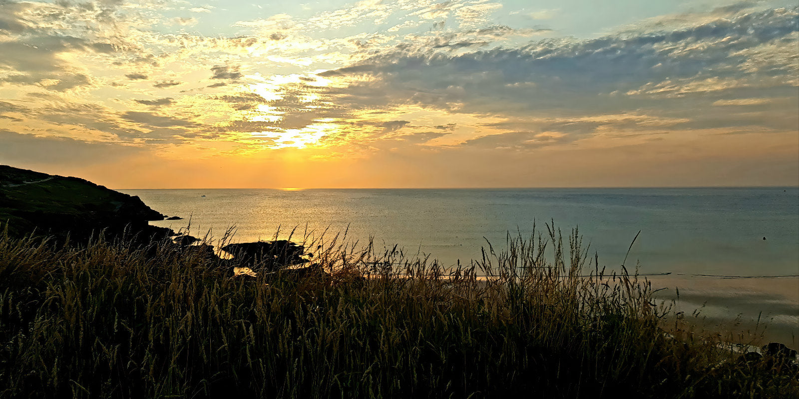 A scenic coastal sunset with the sun low over the horizon, glowing yellow light reflecting on calm water, and dark grasses silhouetted in the foreground.