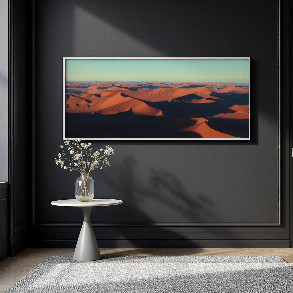 A framed landscape art print depicting orange sand dunes under a pale sky, displayed on a dark charcoal wall above a small white side table with a vase of white flowers.