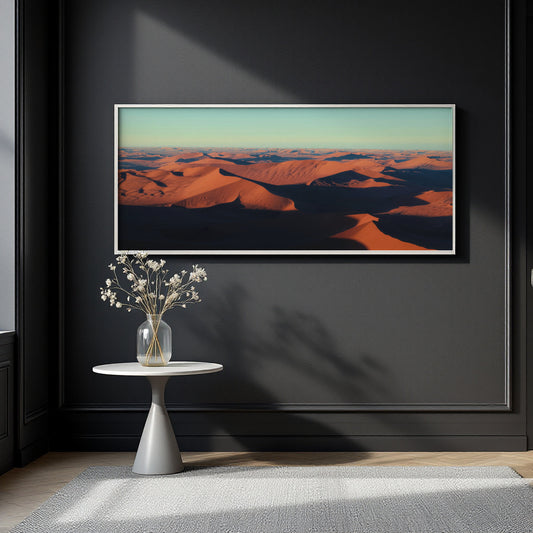 A framed landscape art print depicting orange sand dunes under a pale sky, displayed on a dark charcoal wall above a small white side table with a vase of white flowers.