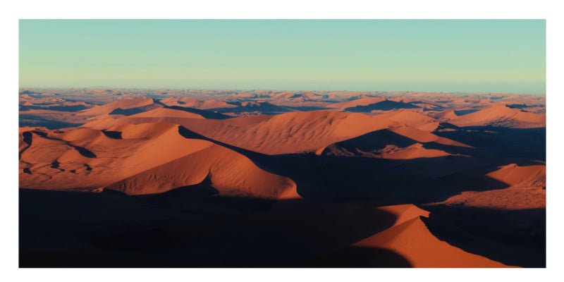 A scenic landscape photograph of rolling desert sand dunes in warm orange-brown tones under a pale blue sky.