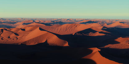Aerial view of vast orange-red sand dunes under a clear pale blue sky, with long shadows stretching across the desert landscape.