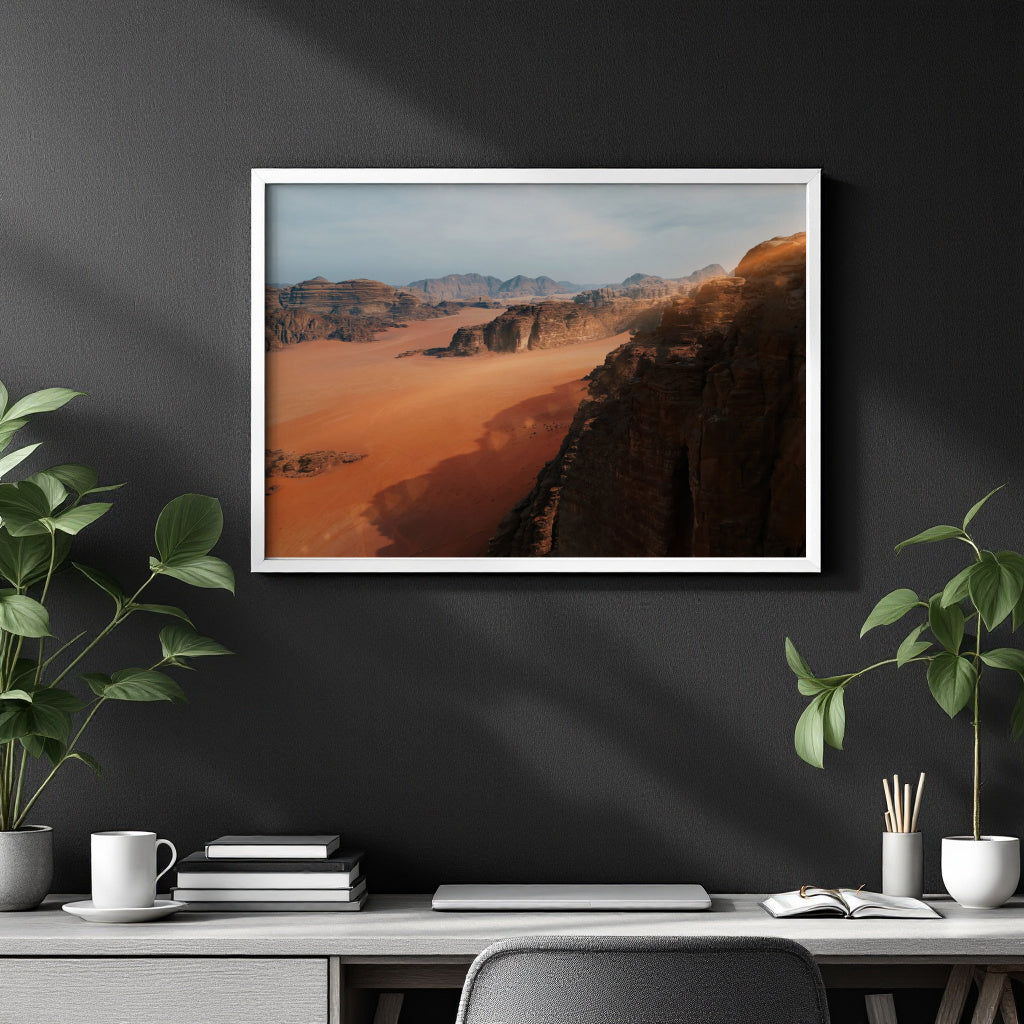 A framed landscape art print of a desert canyon with orange sand and dark rocky cliffs, displayed on a dark gray wall above a styled desk with plants and books.