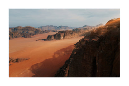 A scenic landscape photograph of a desert canyon with orange sand, dark rock cliffs, and distant mountains under a pale sky, shown with a white border.