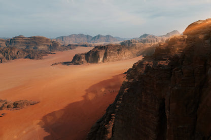 A scenic desert landscape with reddish-orange sand dunes, rugged brown rock formations, and distant mountains under a pale blue sky.