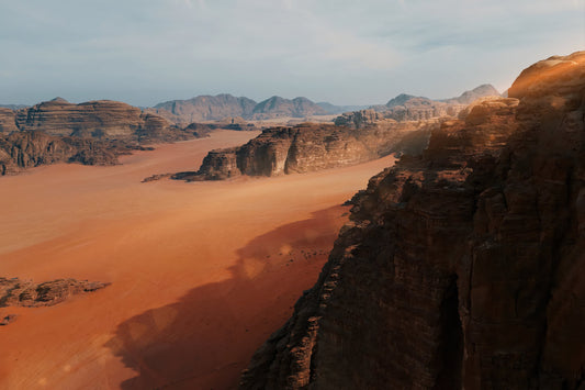 A scenic desert landscape with reddish-orange sand dunes, rugged brown rock formations, and distant mountains under a pale blue sky.