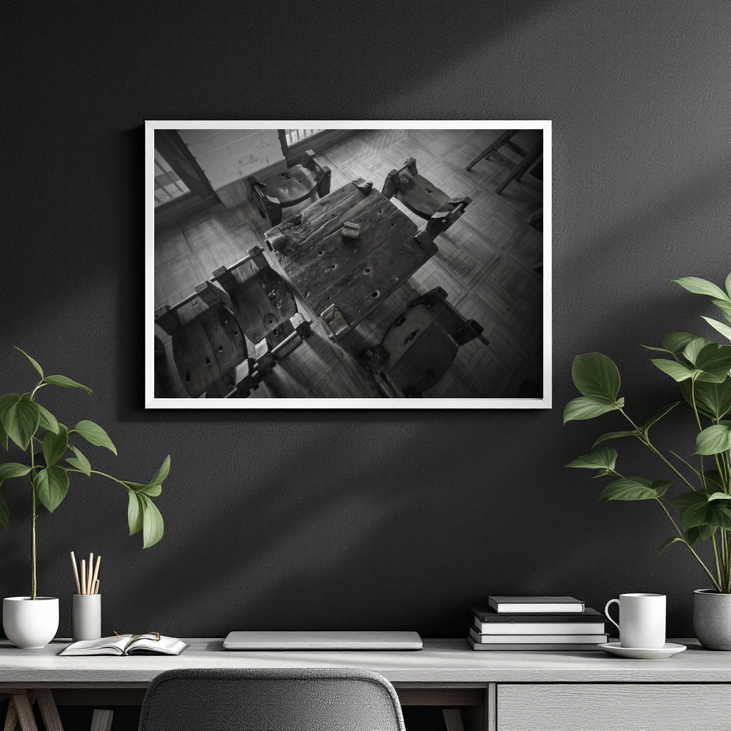 A framed black and white art print depicting a high-angle view of a rustic wooden table and chairs, displayed on a dark gray wall above a styled desk with plants and books.