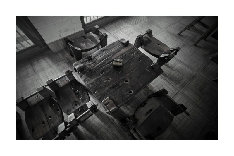 Black and white photograph of a rustic wooden table surrounded by several worn chairs in an indoor room with tiled flooring.