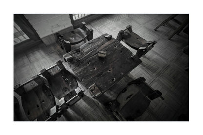 Black and white photograph of a rustic wooden table surrounded by several worn chairs in an indoor room with tiled flooring.