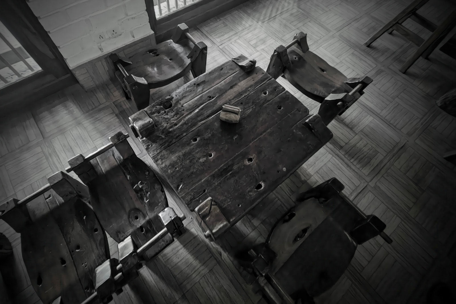 A high-angle black and white photograph of a rustic wooden table surrounded by several worn wooden chairs in a dimly lit room with tiled flooring.