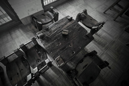 A high-angle black and white photograph of a rustic wooden table surrounded by several worn wooden chairs in a dimly lit room with tiled flooring.