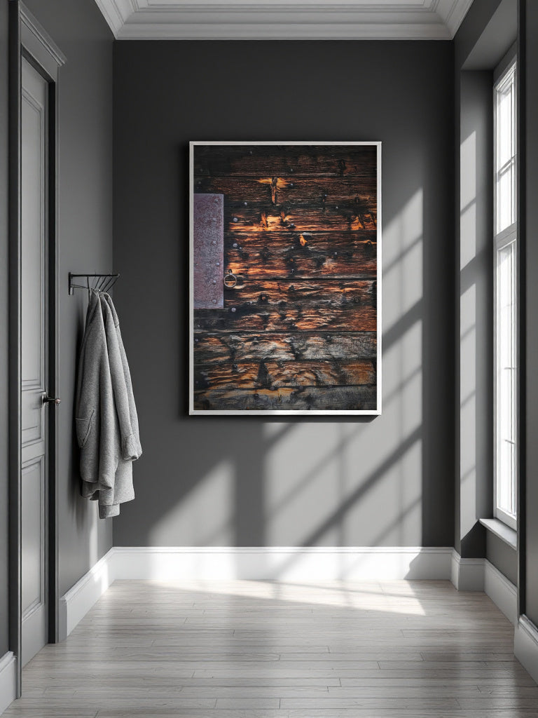 A framed abstract art print featuring textured dark wood grain with warm brown, orange, and black tones, hanging on a dark gray wall in a modern hallway with light-colored wood floors and a window casting shadows.