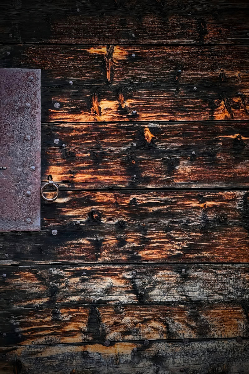 Close-up of a rustic dark wooden door with weathered planks, visible grain, nail heads, and a metal knocker on the left.