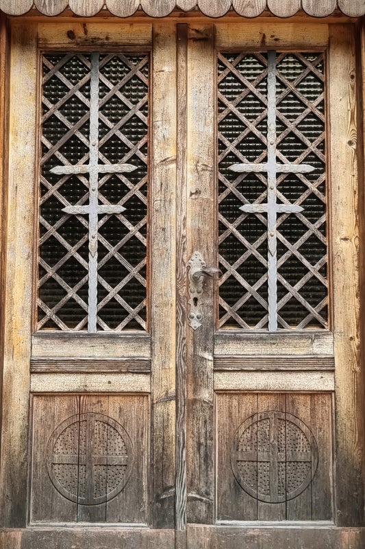 Close-up of weathered wooden double doors with diamond-pattern metal grilles in the upper windows and circular carved details on the lower panels.
