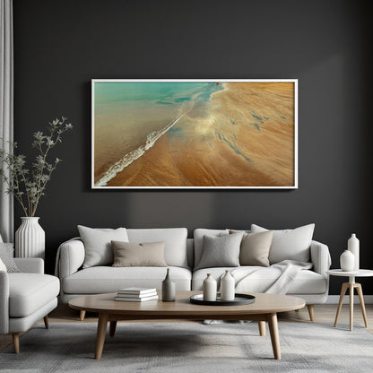 A framed landscape art print depicting a sandy beach meeting the ocean, displayed on a dark gray wall above a modern white sofa in a living room setting.