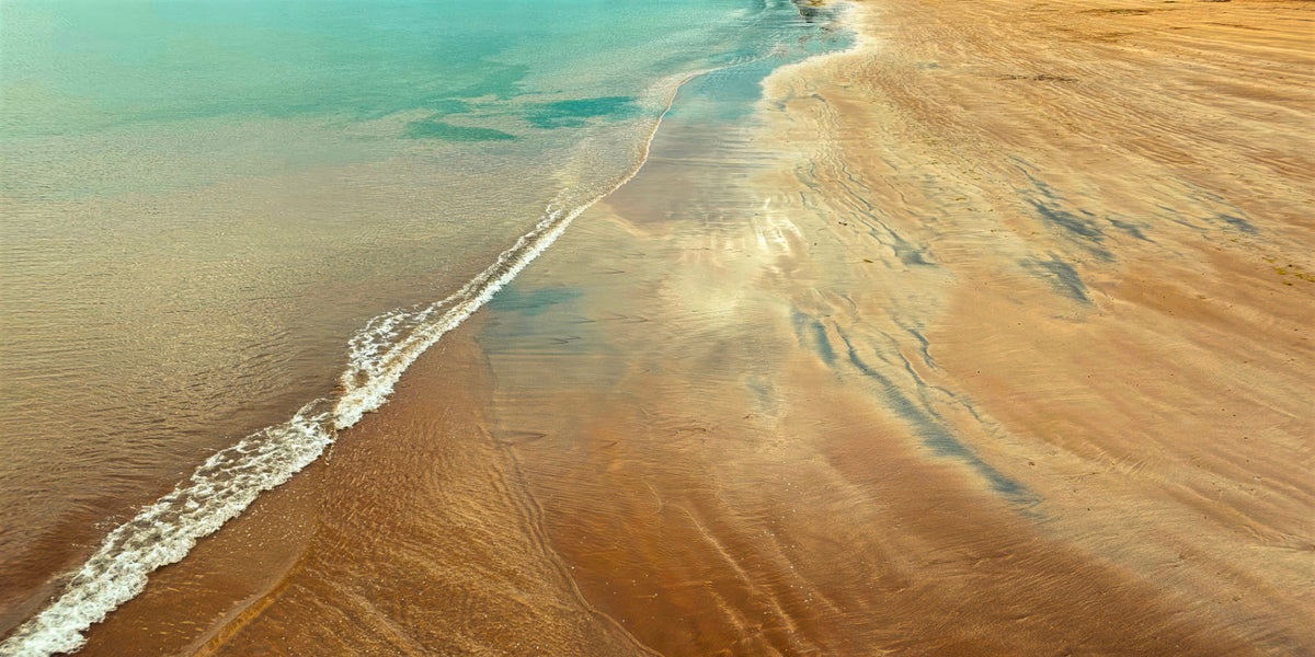 Aerial view of a sandy beach shoreline with turquoise water and pale brown sand, showing ripple patterns and a white line of small waves.