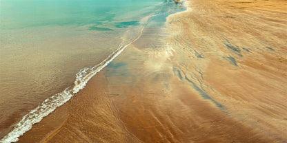 Aerial view of a sandy beach shoreline with turquoise water and pale brown sand, showing ripple patterns and a white line of small waves.