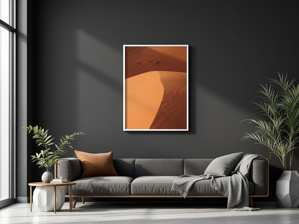 A framed vertical art print of desert sand dunes in warm orange and brown tones, displayed on a dark charcoal wall above a modern gray sofa in a styled living room.