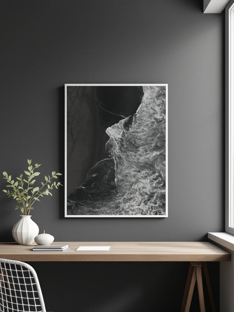 A framed black and white abstract art of ocean waves print hanging on a dark gray wall above a wooden desk with a vase and plant.