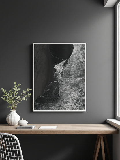 A framed black and white abstract art of ocean waves print hanging on a dark gray wall above a wooden desk with a vase and plant.