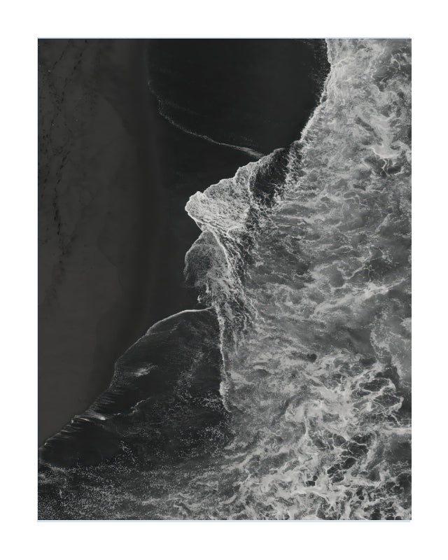 A black and white aerial photograph of a coastline, showing the edge of a sandy beach meeting foamy ocean waves, presented as a square print with a white border.