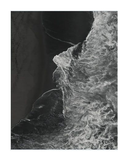 A black and white aerial photograph of a coastline, showing the edge of a sandy beach meeting foamy ocean waves, presented as a square print with a white border.