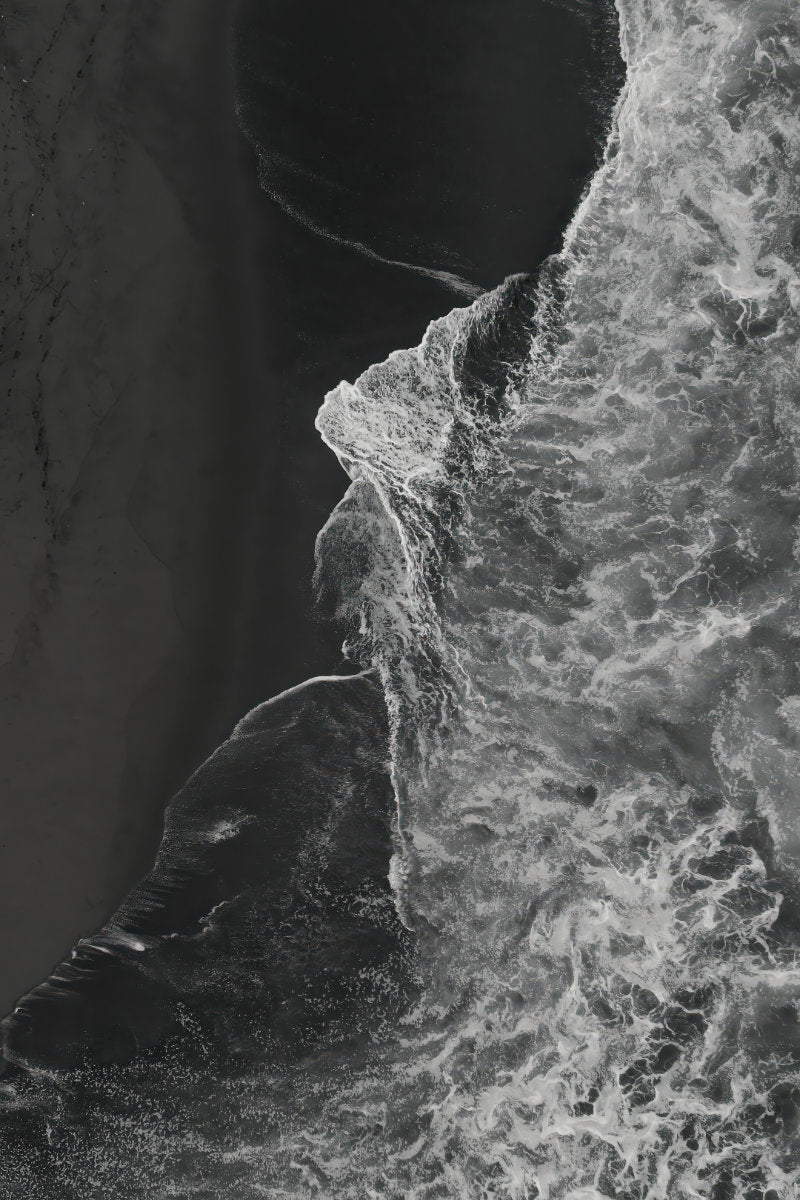 Black and white aerial photograph of ocean waves crashing against a dark rocky shoreline, with high-contrast textures of foam and sand.